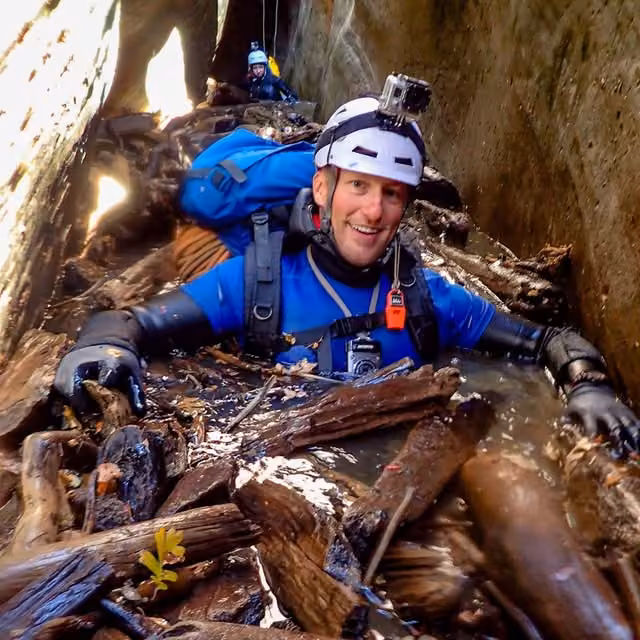 A smiling canyoneer in blue gear and a white helmet in a water-filled rocky passage